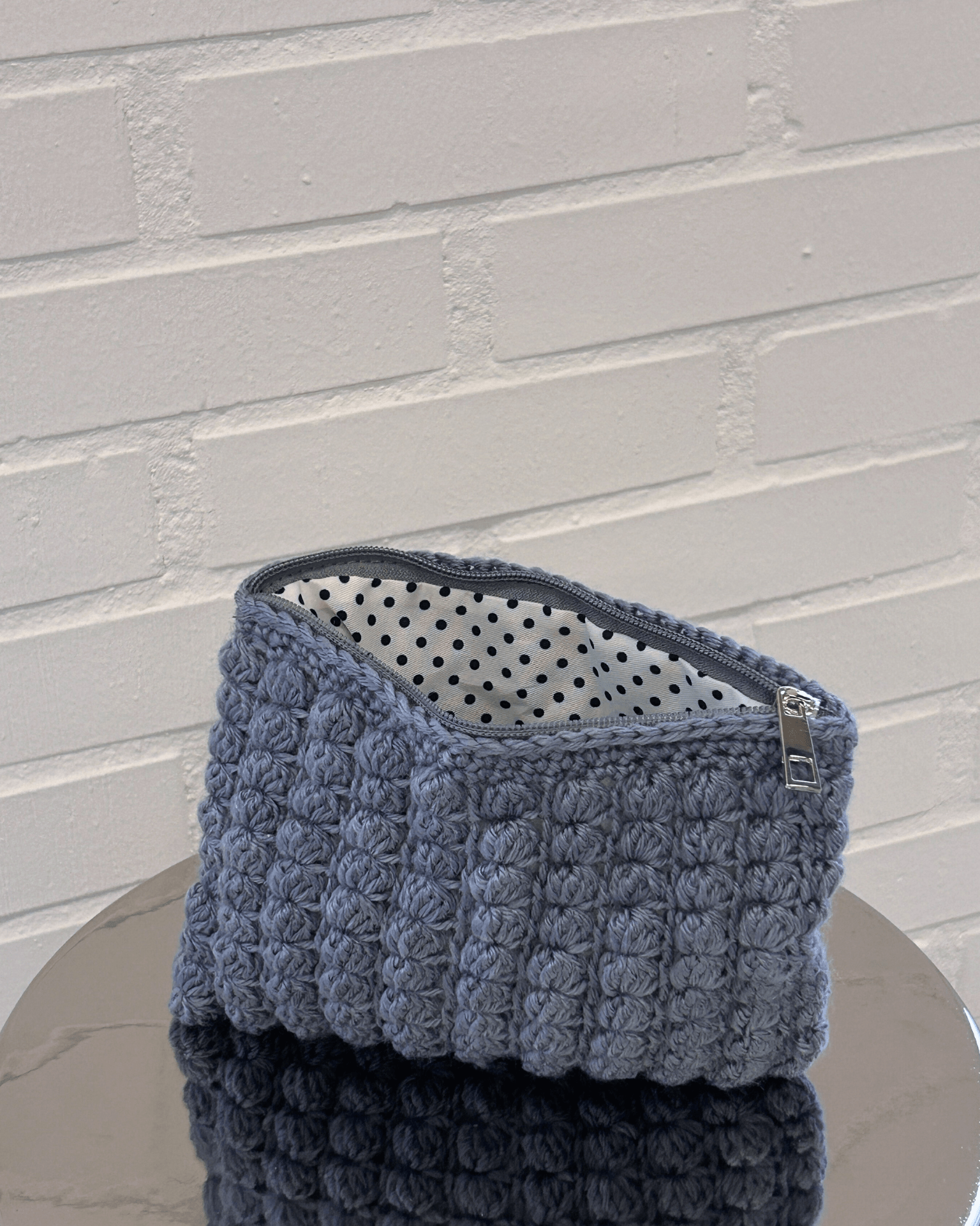 Grå hæklet clutch med prikket foer og lynlås på et spejl