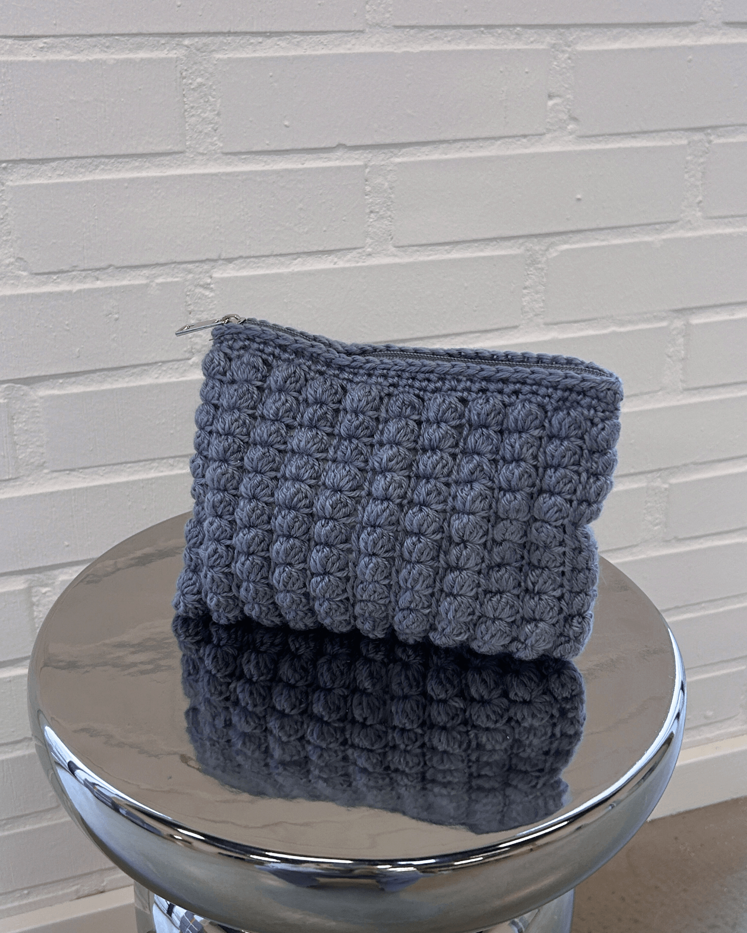 Hæklet grå clutch i boblemønster med lynlås på en metalskammel