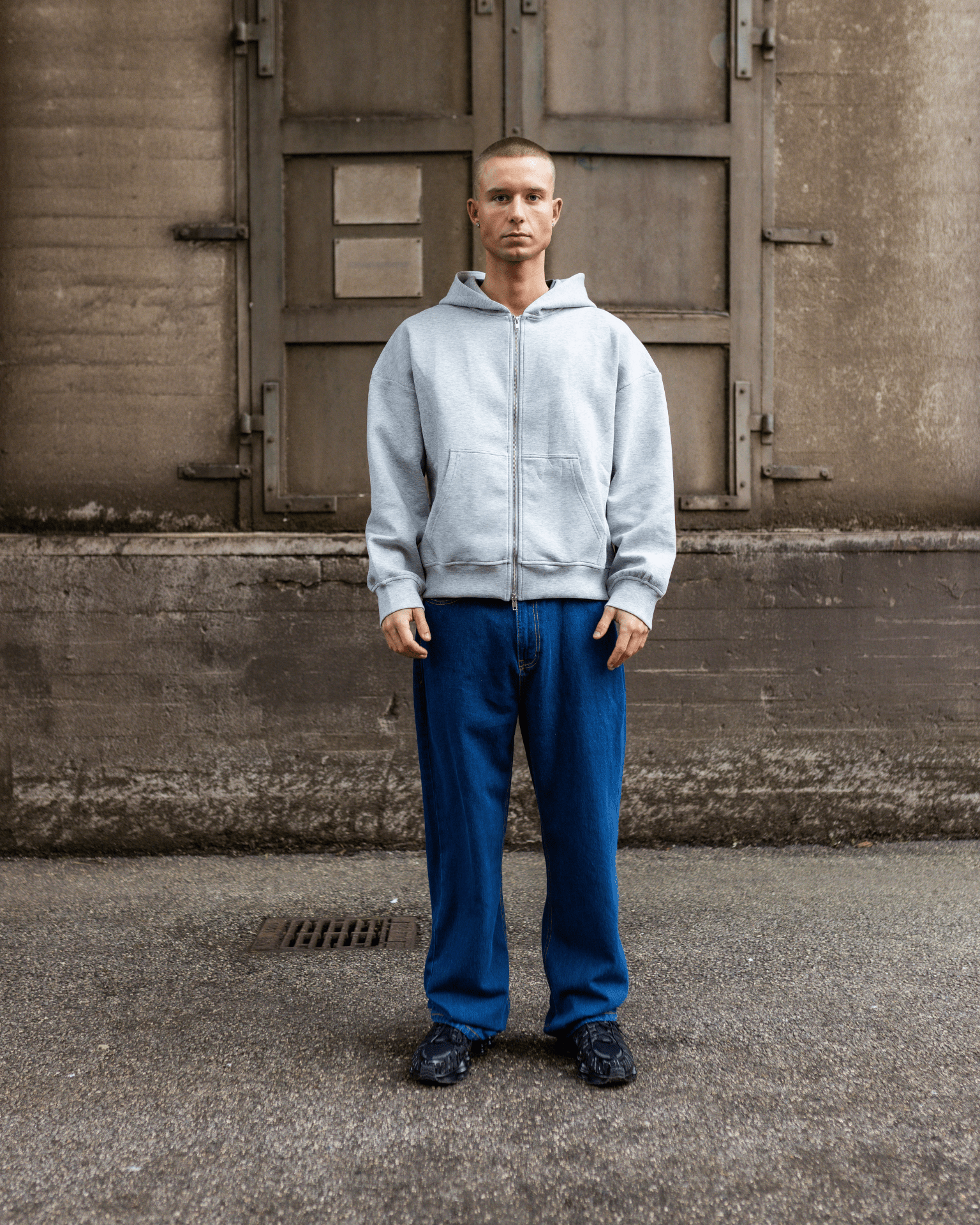 Mand i mørkeblå Tyler baggy jeans og grå hættetrøje med lynlås.