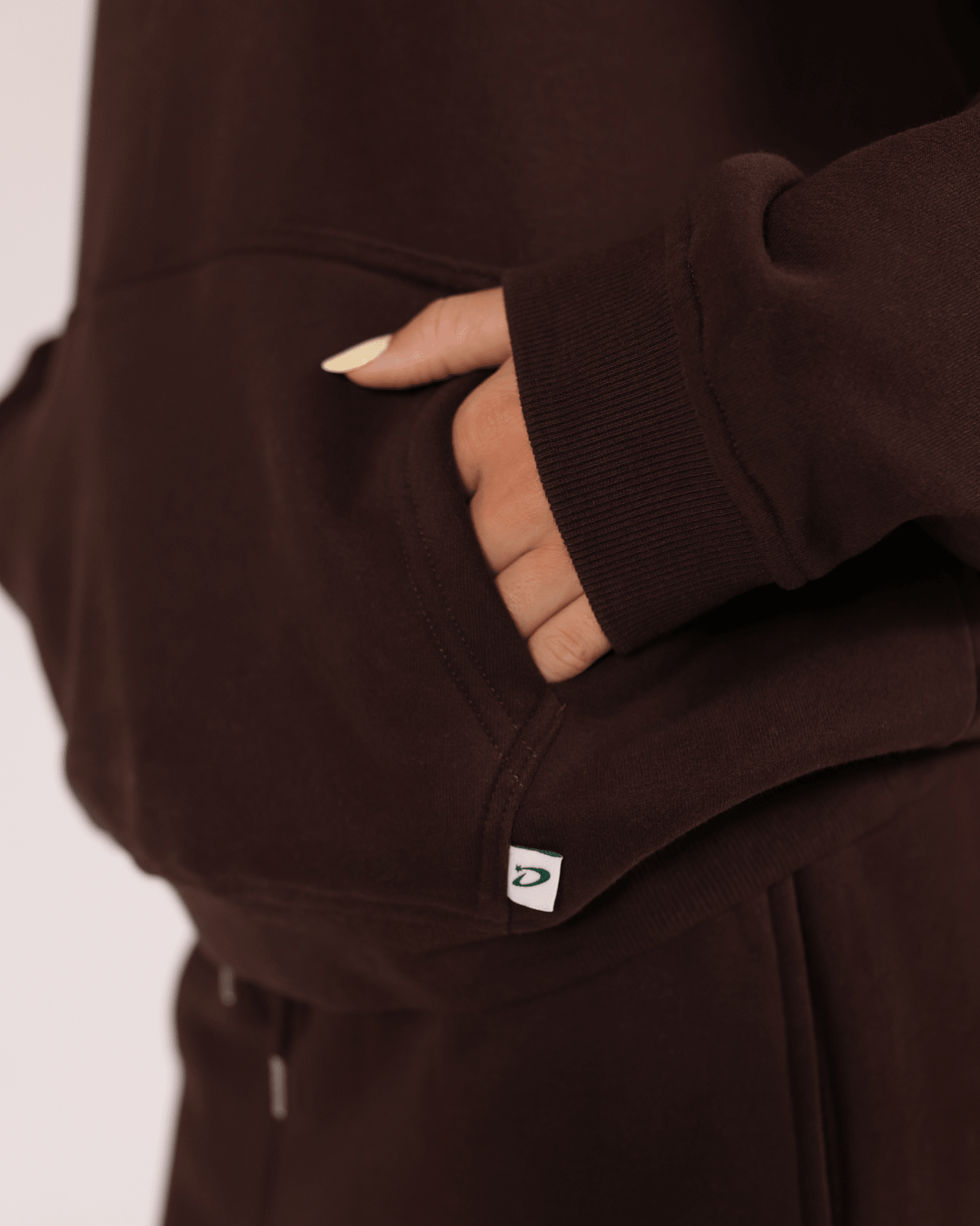 Nærbillede af brun oversized bomuldshoodie med kængurulomme og lille hvidt logo.